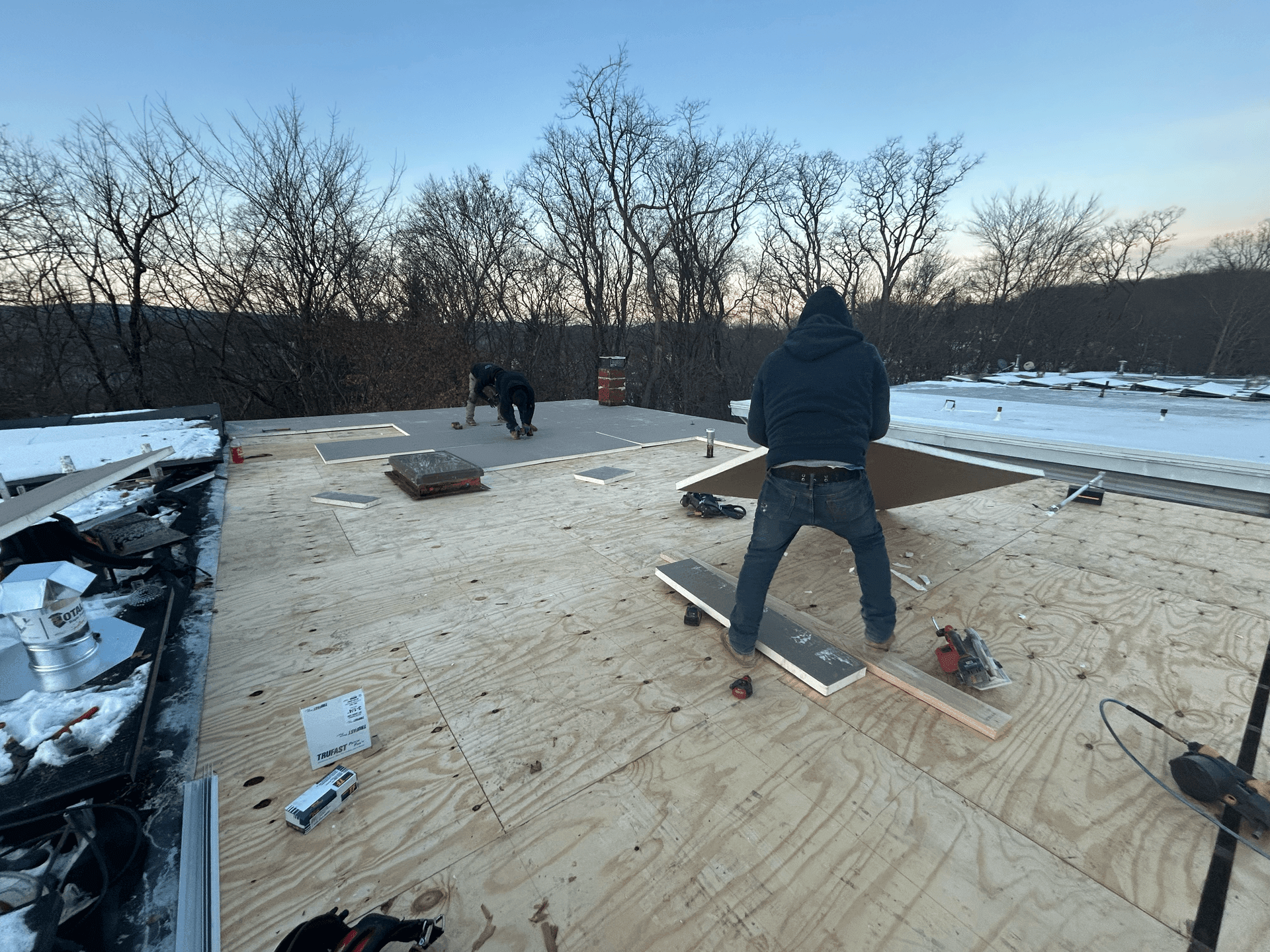 Two workers installing plywood boards on a rooftop with bare trees and a winter landscape in the background.